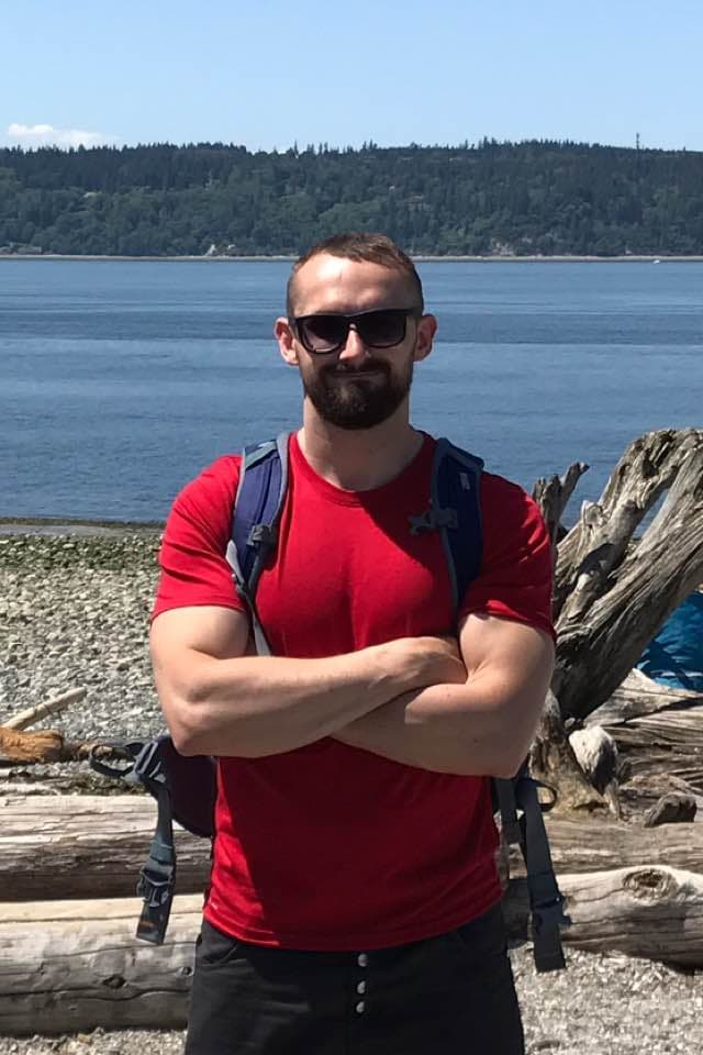 A man with a beard and sunglasses standing on a rocky beach with a forested background.